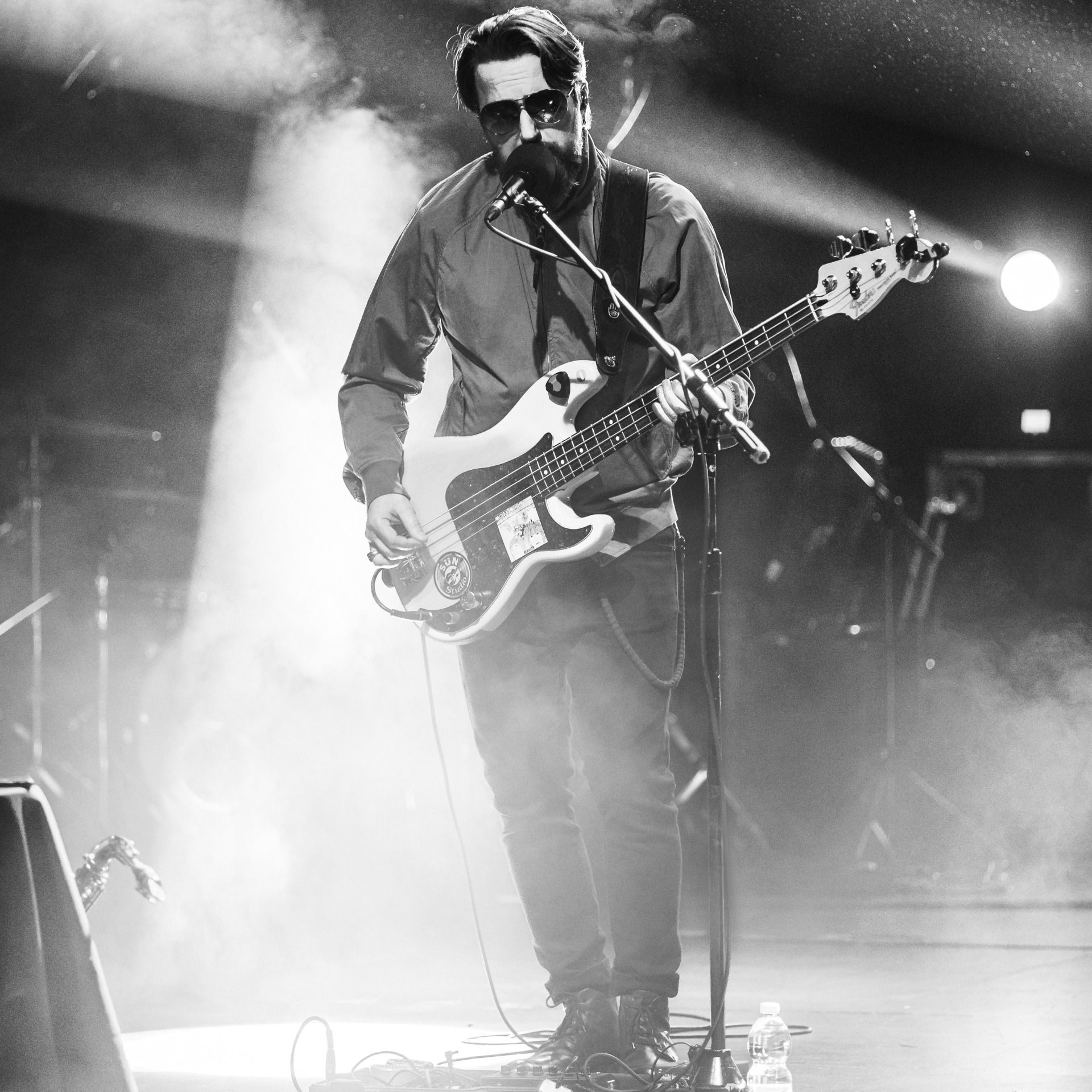 Czarno-biała fotografia koncertowa przedstawia muzyka z brodą i w okularach przeciwsłonecznych grającego na gitarze basowej podczas występu na scenie w oświetleniu scenicznym i lekkiej mgle.