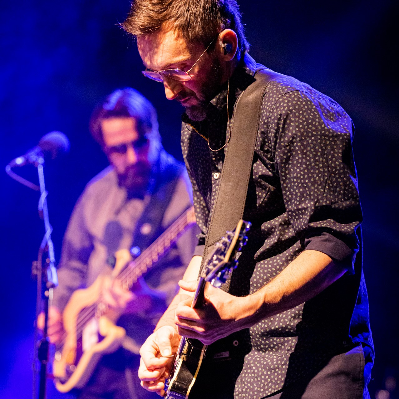 Gitarzysta z brodą i okularami gra na czarnej gitarze na scenie oświetlonej niebieskim światłem, w tle widoczny basista z mikrofonem.