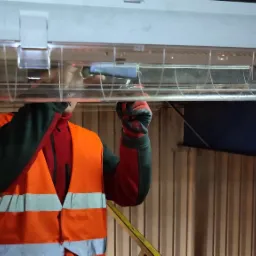 Elektryk w kamizelce odblaskowej montuje podłużną lampę sufitową z przezroczystą obudową, używając narzędzia w rękawicach ochronnych.