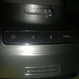 Fragment robota kuchennego KitchenAid z widocznym panelem sterowania, ustawieniami grubości krojenia i logiem marki, umieszczony na wzorzystym obrusie.