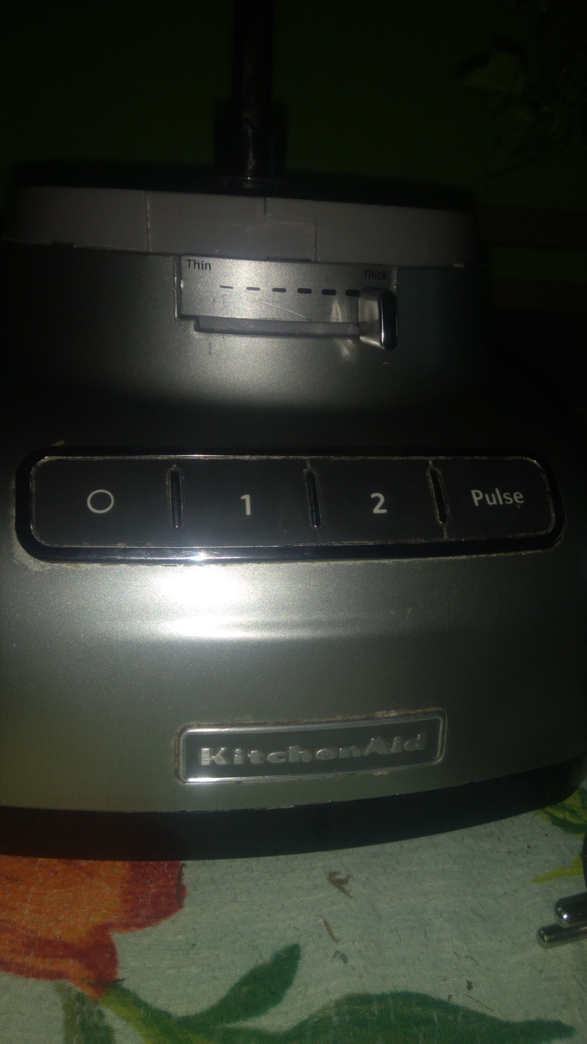 Fragment robota kuchennego KitchenAid z widocznym panelem sterowania, ustawieniami grubości krojenia i logiem marki, umieszczony na wzorzystym obrusie.