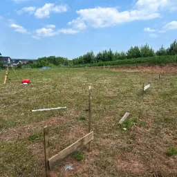 Wytyczenie budynku: tyczka geodezyjna, niwelator i skrzynka na trawiastym terenie budowy przy domu z czarnym dachem i białymi ścianami w słoneczny dzień.