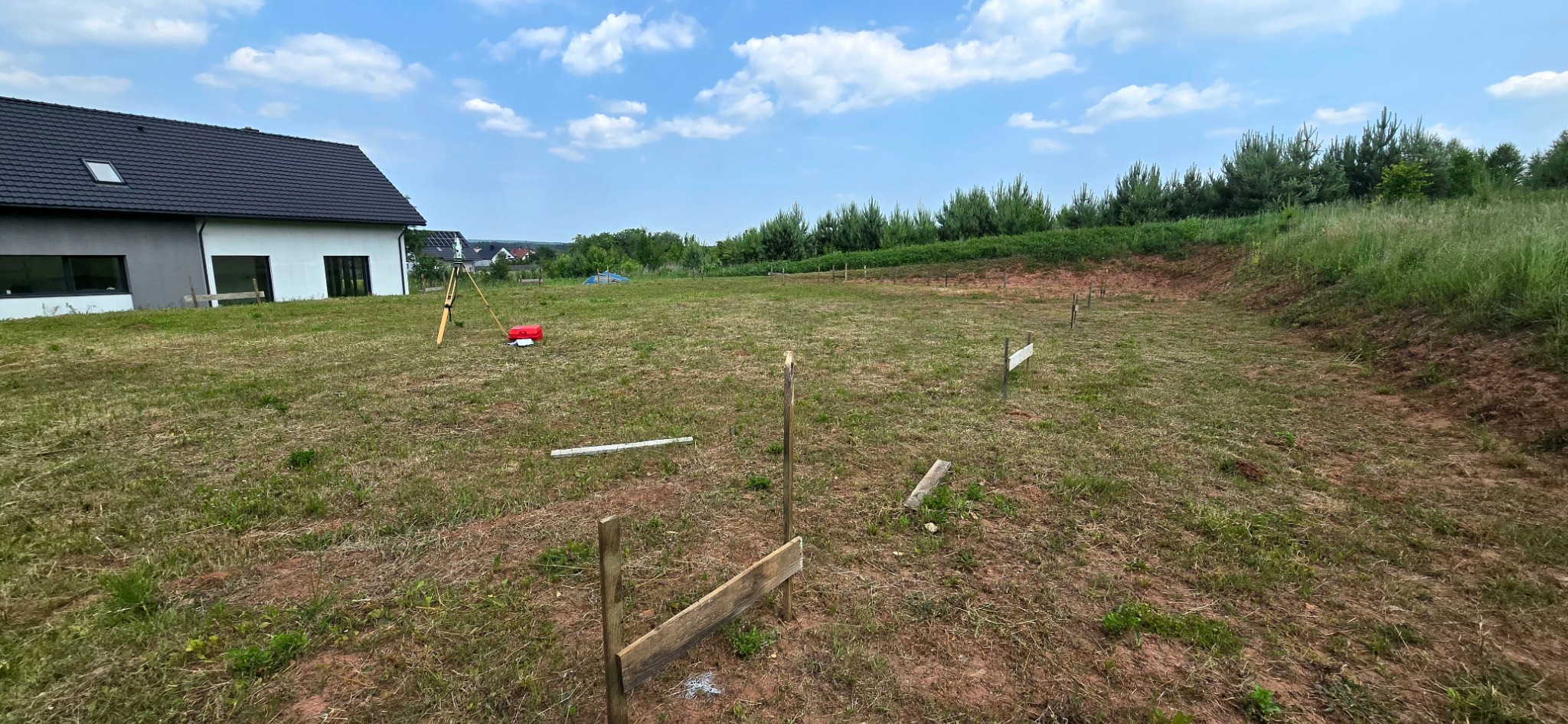 Wytyczenie budynku: tyczka geodezyjna, niwelator i skrzynka na trawiastym terenie budowy przy domu z czarnym dachem i białymi ścianami w słoneczny dzień.