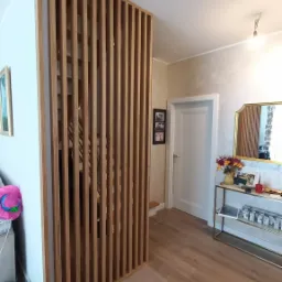 Drewniana ścianka działowa z pionowych listew, oddzielająca schody, w jasnym wnętrzu domu w Ciechanowie. Widoczne schody przez prześwity oraz fragment wyposażenia wnętrza.