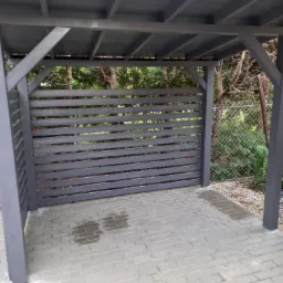 Szara altana z drewnianymi, poziomymi panelami, widok z zewnątrz na brukowaną podłogę z plamami wody i siatkę ogrodzeniową w tle.