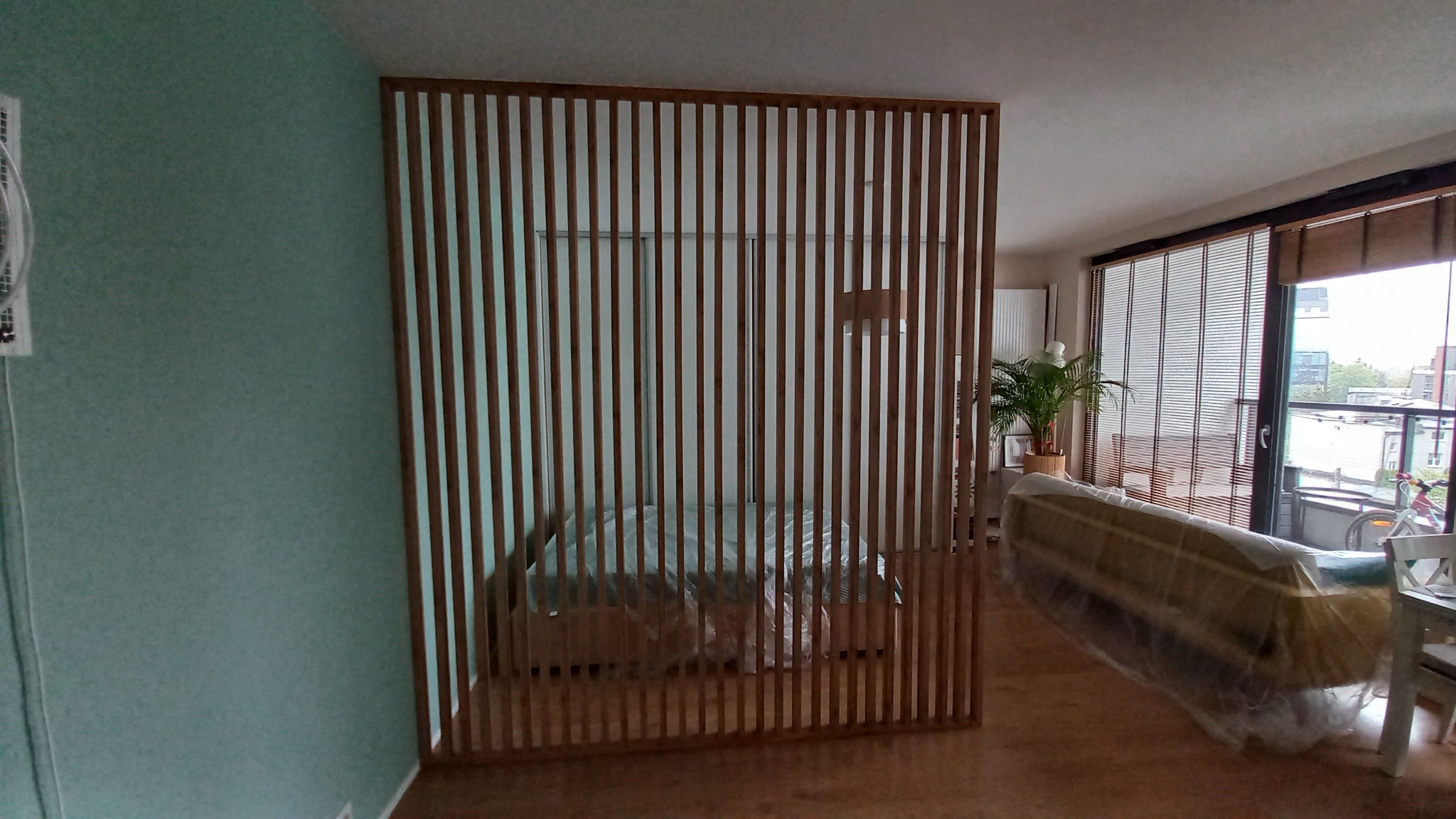 Drewniana ścianka działowa z pionowych listew w jasnym wnętrzu mieszkania, meble zabezpieczone folią, widok na balkon i zieleń za oknem.