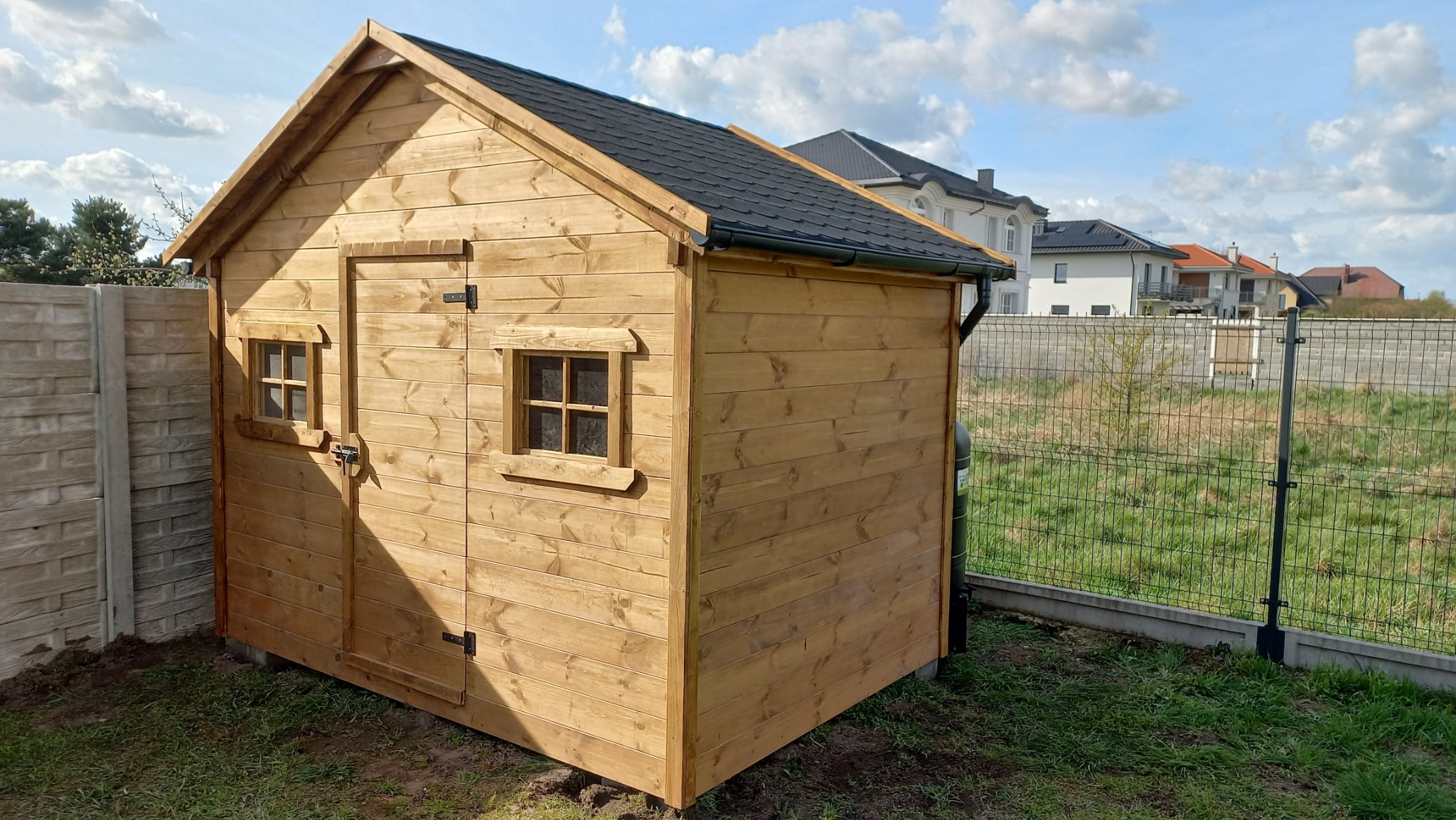 Drewniany domek narzędziowy z czarnym dachem, umieszczony na trawniku w ogrodzie, z widocznym ogrodzeniem panelowym i domami w tle.