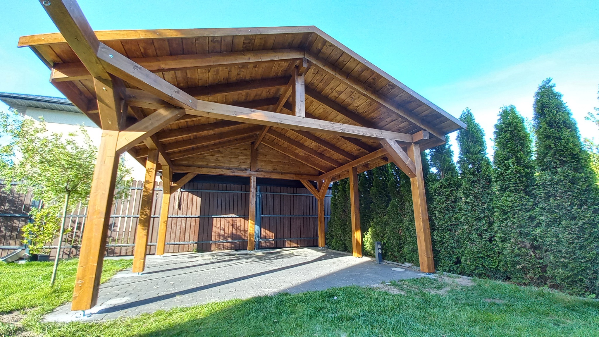 Drewniana konstrukcja z zadaszeniem, prawdopodobnie wiata garażowa lub zadaszenie tarasu, z widocznymi elementami konstrukcyjnymi i betonową nawierzchnią, otoczona zielenią i tujami.