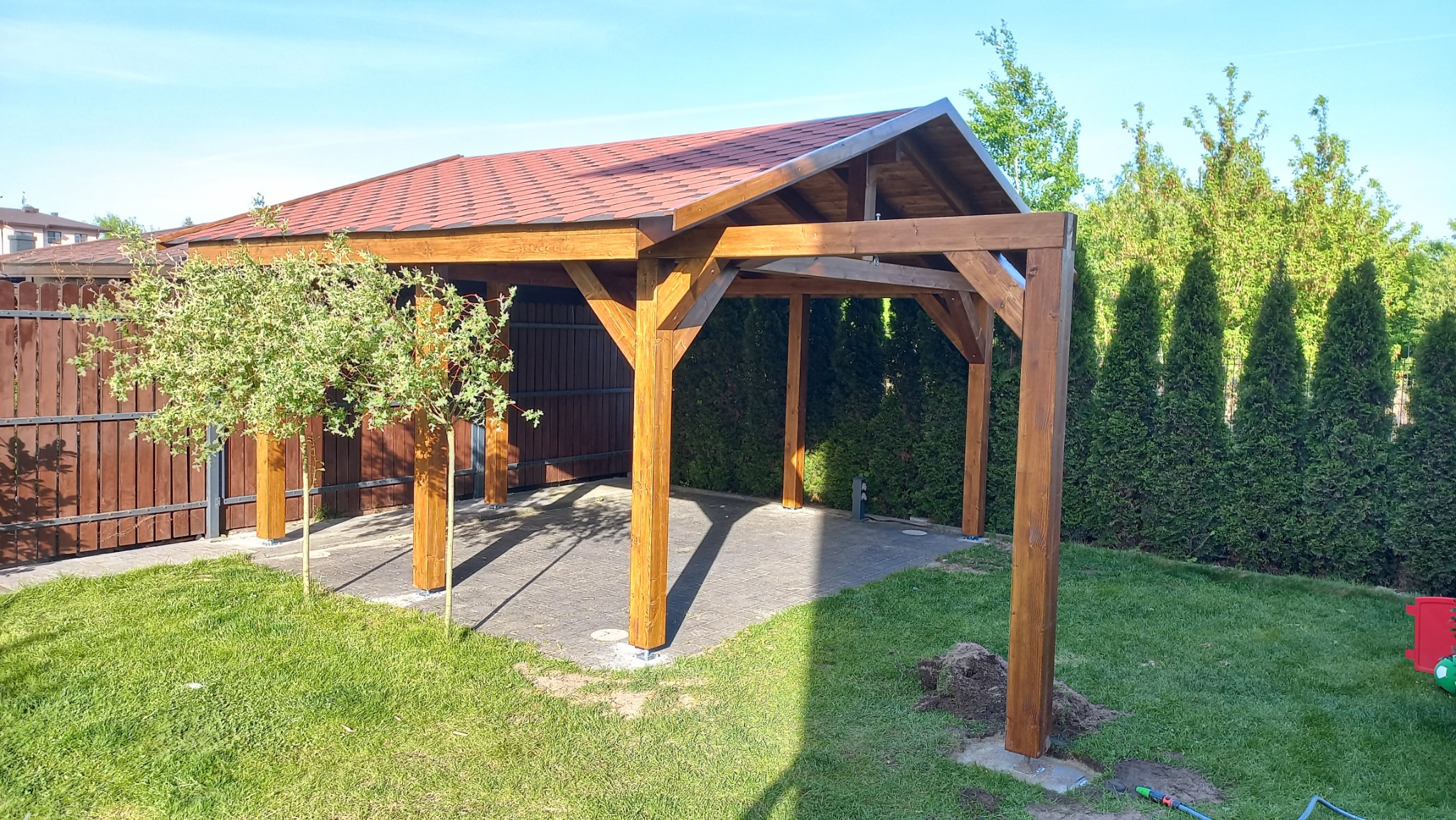 Drewniana altana z czerwoną dachówką na posesji z zielonym trawnikiem i tujami, widoczne świeżo posadzone drzewka liściaste.