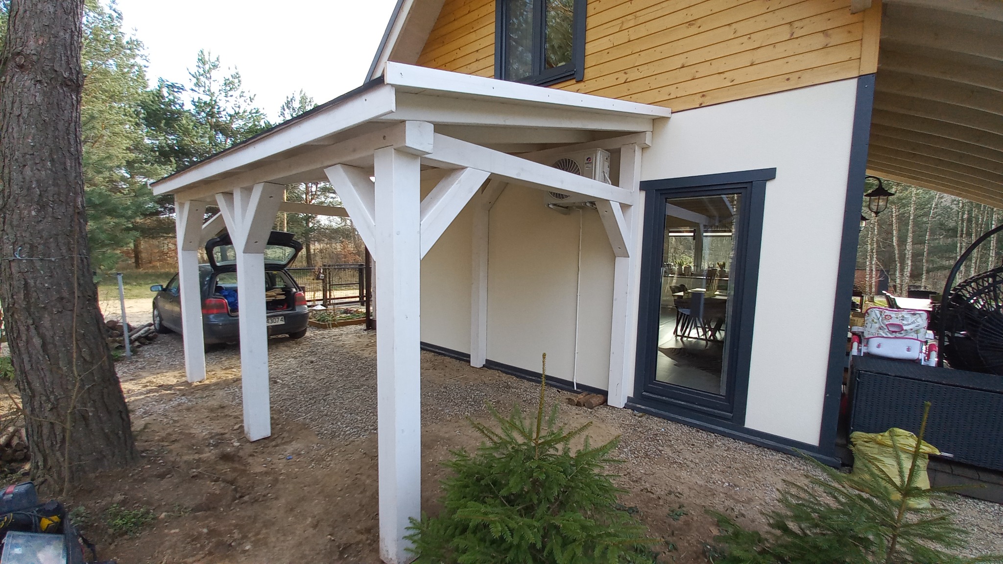 Biała konstrukcja z drewna, prawdopodobnie zadaszenie tarasu lub wejścia, przylegająca do jasnego budynku z drewnianą elewacją, na żwirowym podłożu, z samochodem w tle i małą choinką na pierwszym...