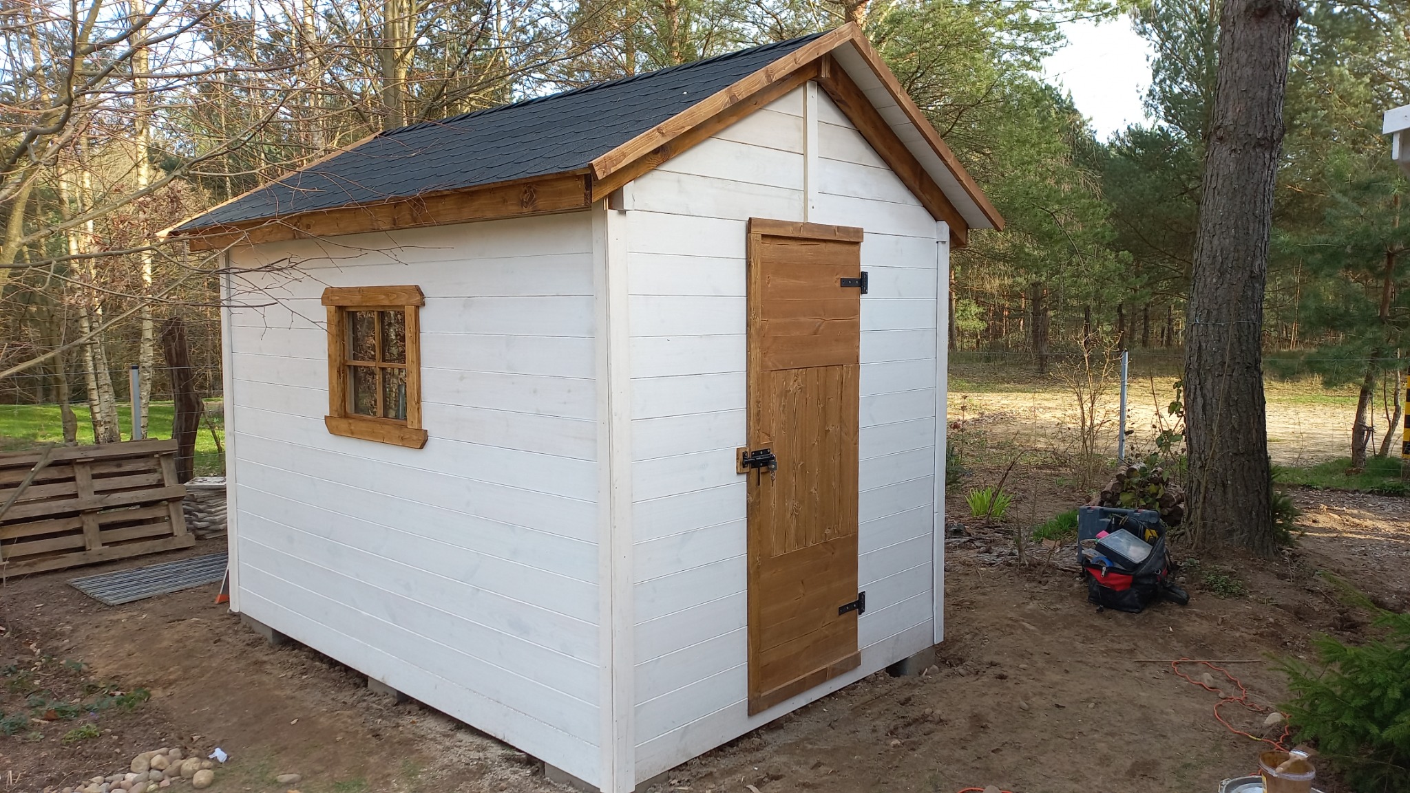 Biała, drewniana konstrukcja z oknem i drzwiami, przypominająca mały domek gospodarczy z ciemnym dachem, otoczona drzewami i narzędziami.