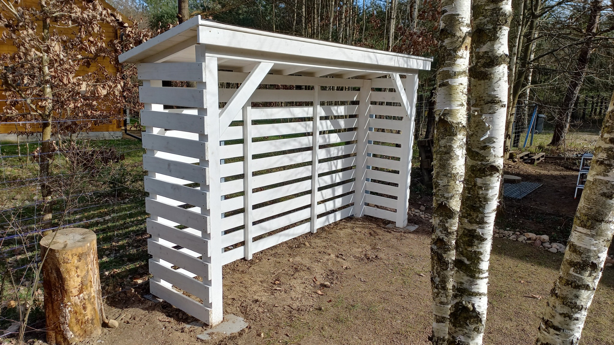 Biała drewniana pergola z poziomymi szczebelkami, widok z zewnątrz, w tle drzewa i siatka ogrodzeniowa.