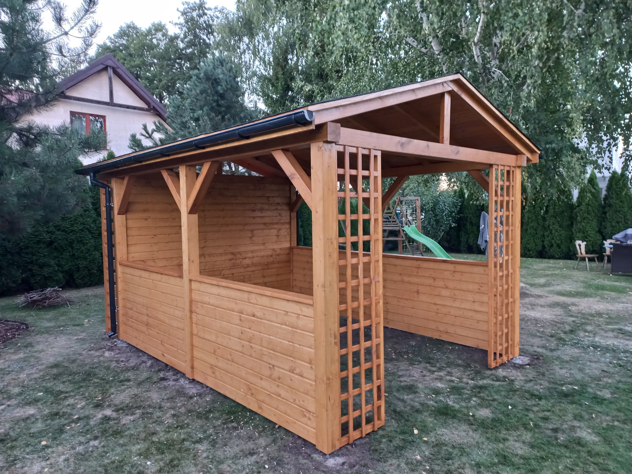 Drewniana konstrukcja z kratownicami, częściowo zabudowana deskami, na trawniku przed domem, z widocznym fragmentem zjeżdżalni w tle.