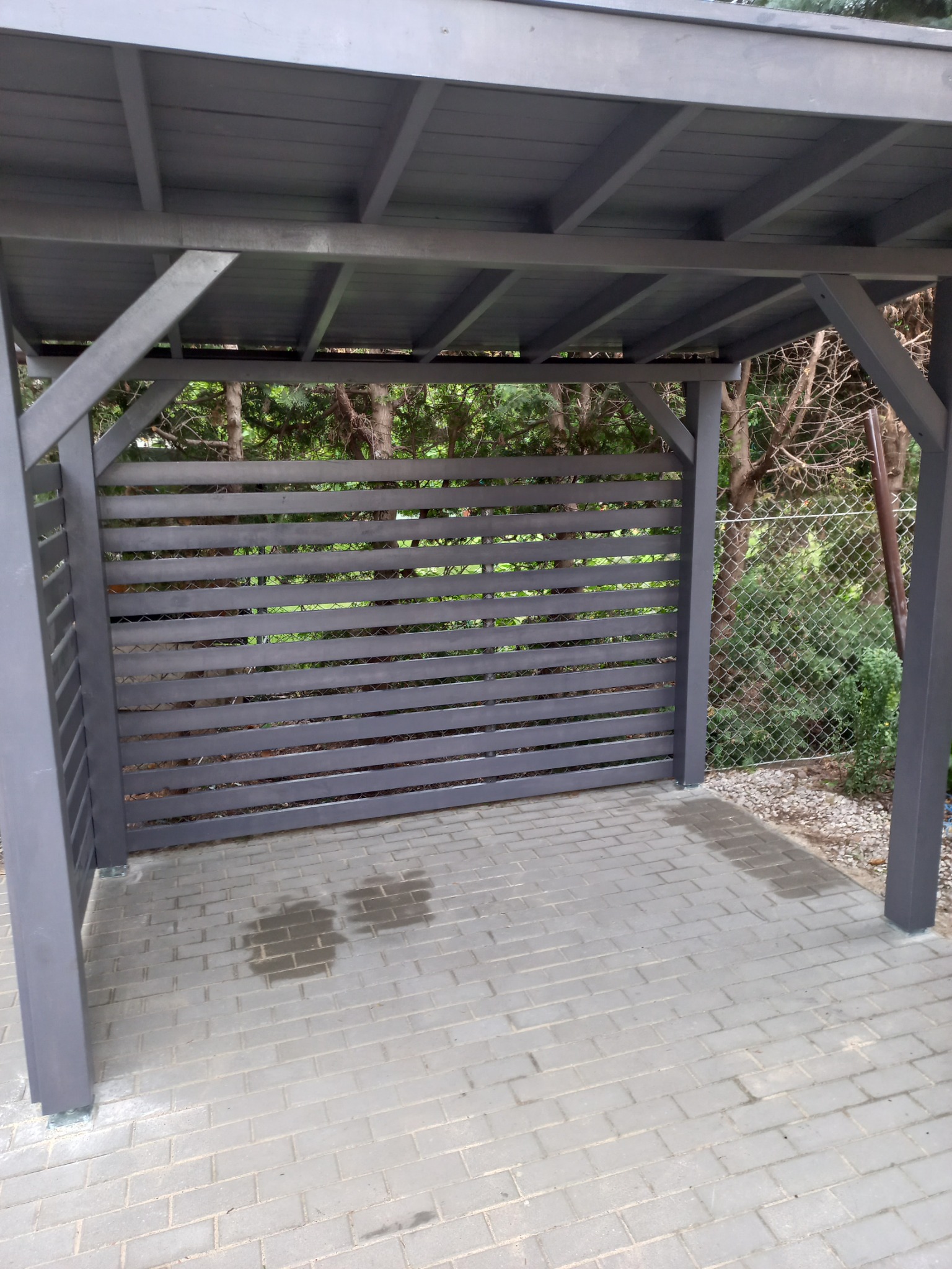 Szara altana z drewnianymi, poziomymi panelami, widok z zewnątrz na brukowaną podłogę z plamami wody i siatkę ogrodzeniową w tle.