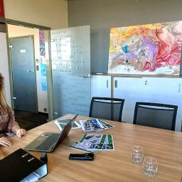 Lokalne Nieruchomości Agnieszka Grzelewska - Kobieta siedzi za stołem w biurze z laptopem i dokumentami. W tle obrazy, szklane drzwi z napisami i szafki.