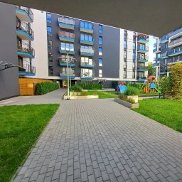 Lokalne Nieruchomości Agnieszka Grzelewska - Nowoczesny kompleks apartamentów z placem zabaw i zadbaną zielenią, widok z perspektywy chodnika z kostki brukowej. Jasne, popołudniowe światło.