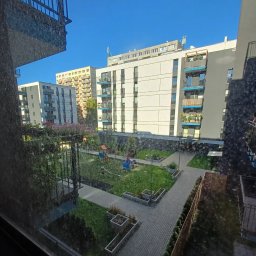 Lokalne Nieruchomości Agnieszka Grzelewska - Widok z okna na osiedle z placem zabaw i zielenią. Budynki z balkonami w tle, błękitne niebo. Ujęcie z wnętrza mieszkania.