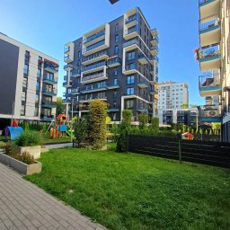 Lokalne Nieruchomości Agnieszka Grzelewska - Nowoczesne osiedle z placem zabaw: wielopiętrowe budynki w odcieniach szarości i bieli z niebieskimi akcentami balkonów, zielony trawnik i kolorowy plac zabaw.