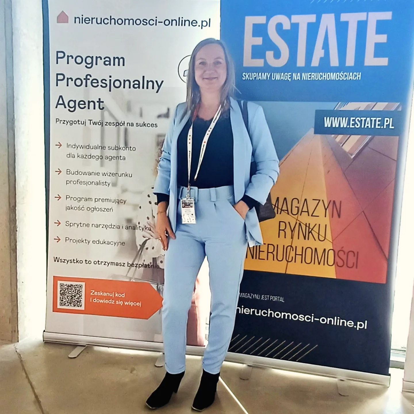 Kobieta w błękitnym garniturze pozuje na tle roll-upu z hasłem 'Program Profesjonalny Agent' i logo 'ESTATE - Magazyn Rynku Nieruchomości'.