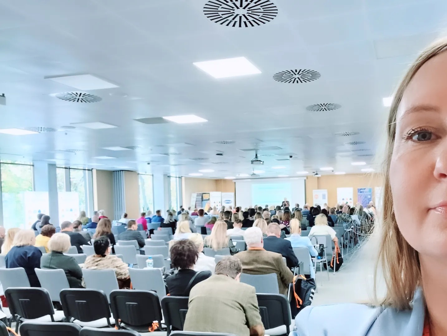 Sala konferencyjna wypełniona uczestnikami podczas prezentacji. Kobieta w jasnej marynarce robi selfie na tle prelekcji. Widoczny projektor i ekrany.