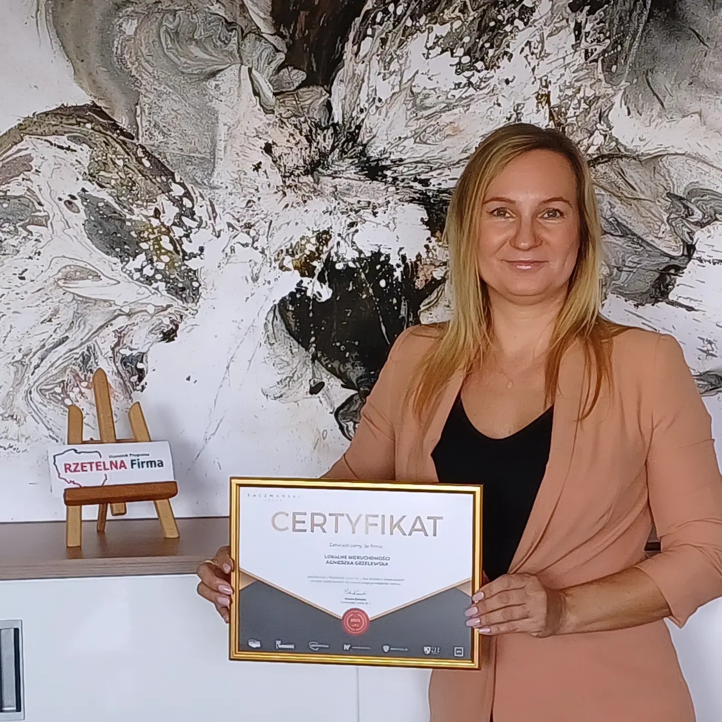 Kobieta prezentuje certyfikat 'Rzetelna Firma' z nazwą 'Lokalne Nieruchomości Agnieszka Gzelewską' na tle abstrakcyjnego obrazu i ekspozytora.