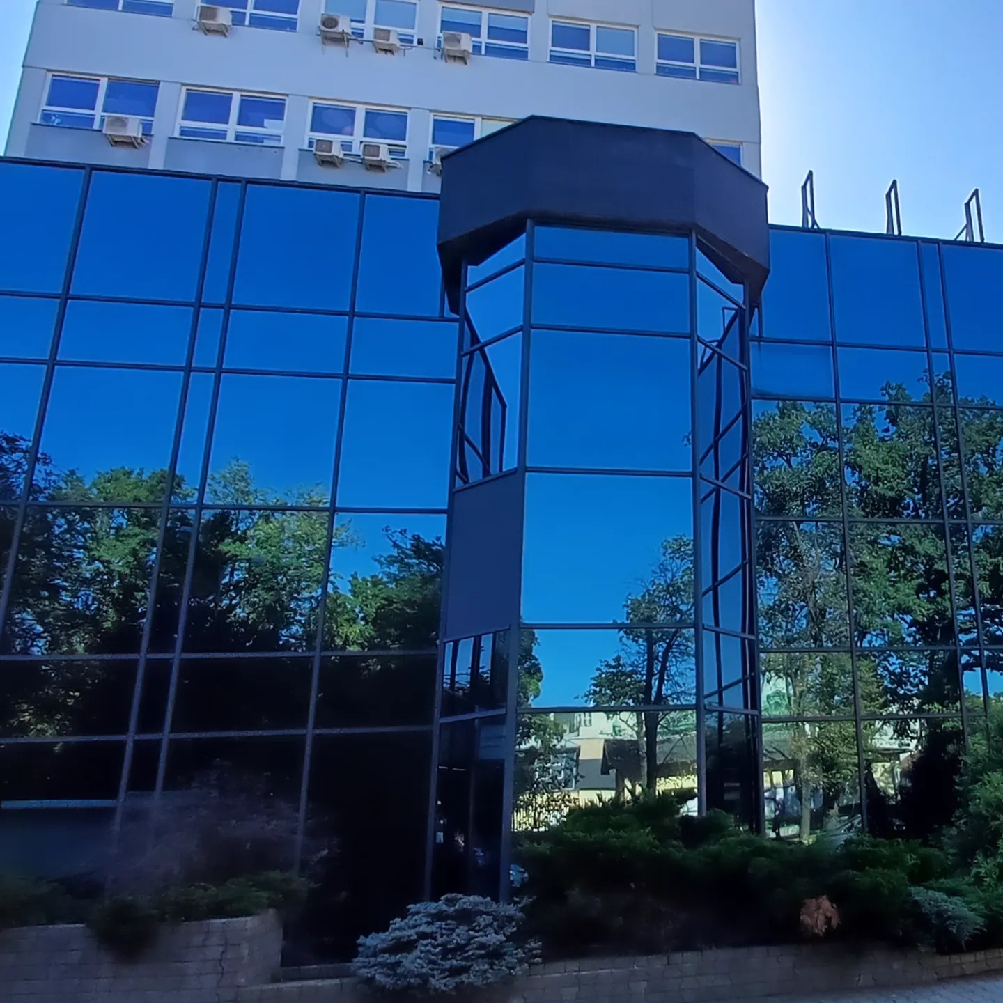 Elewacja biurowca z niebieskimi szybami, w których odbija się zieleń drzew i błękit nieba. Widoczne klimatyzatory na sąsiednim budynku i krzewy przed budynkiem.