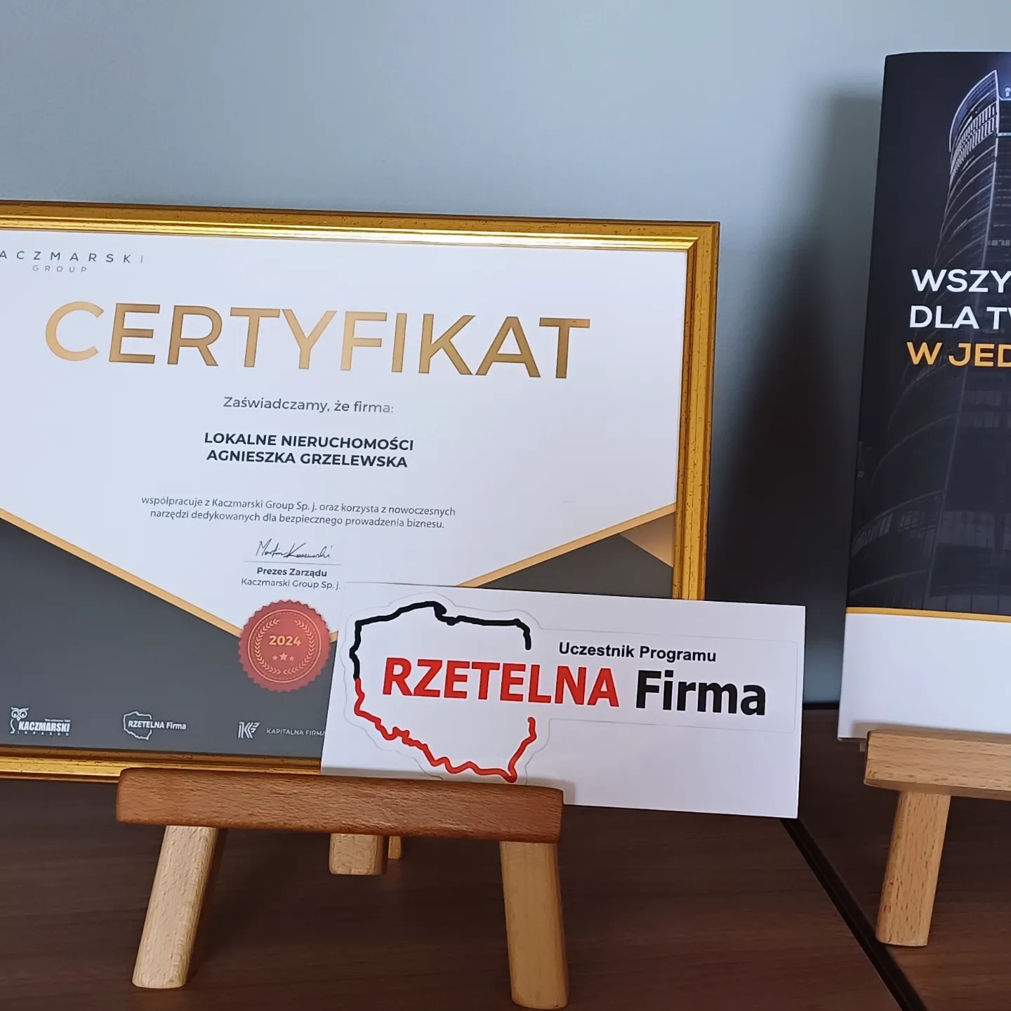 Certyfikat 'Rzetelna Firma' dla Lokalne Nieruchomości Agnieszka Grzelewska, wystawiony przez Kaczmarski Group Sp. j., umieszczony na drewnianym stojaku.