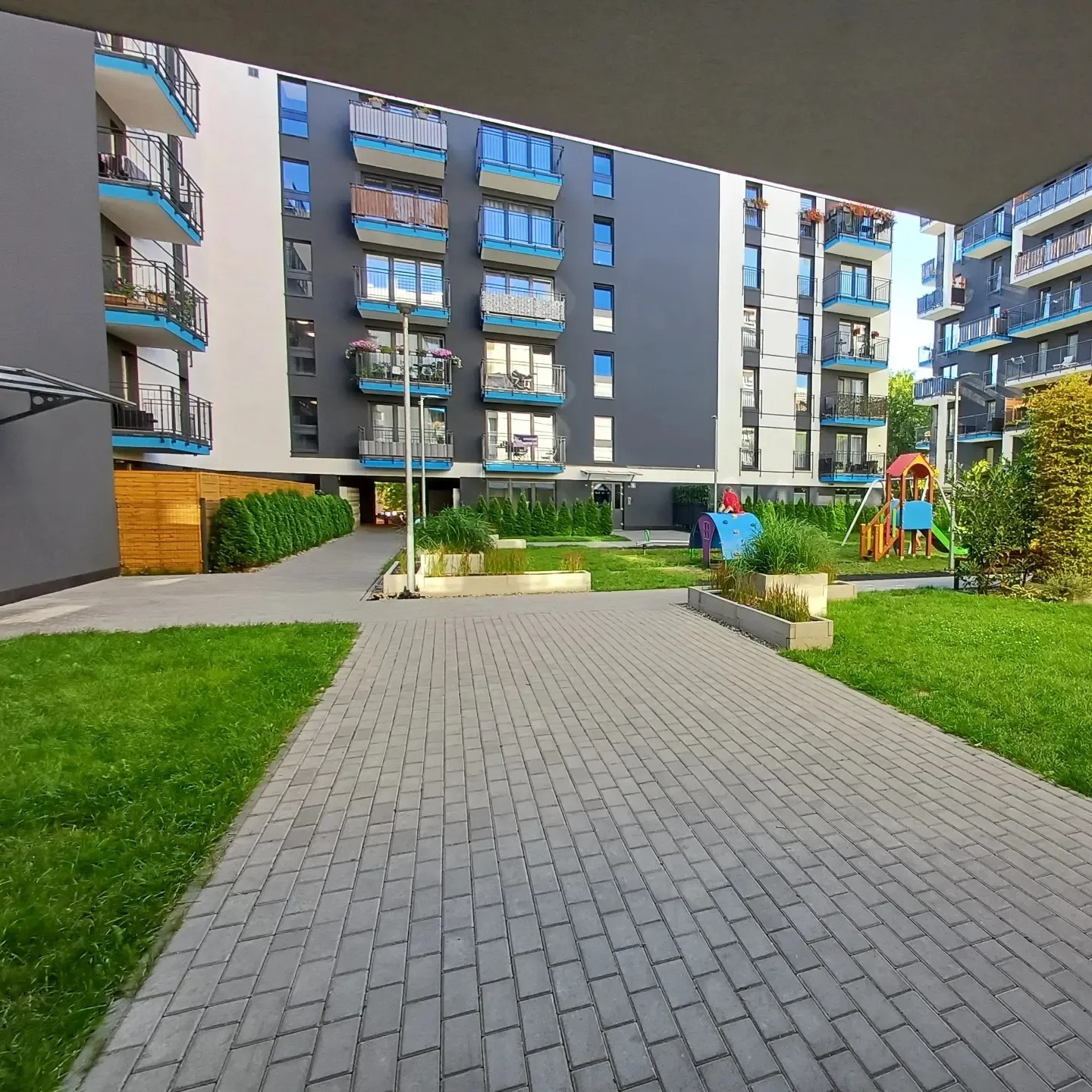 Nowoczesny kompleks apartamentów z placem zabaw i zadbaną zielenią, widok z perspektywy chodnika z kostki brukowej. Jasne, popołudniowe światło.