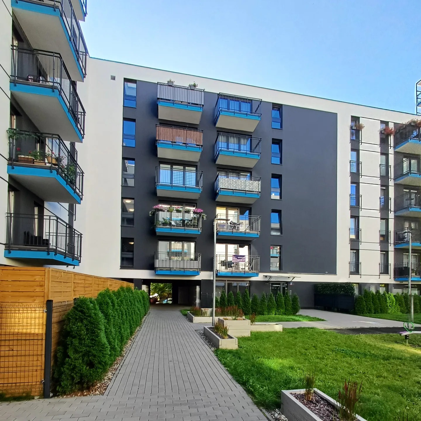 Nowoczesny budynek wielorodzinny z balkonami o niebieskich akcentach, otoczony zielenią i brukowanym chodnikiem w słoneczny dzień.