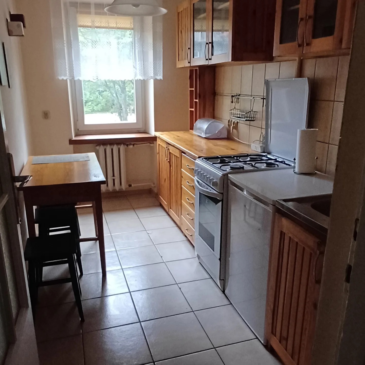 Jasna kuchnia z drewnianymi szafkami, białym piecem i stołem z krzesłami. Okno z widokiem na zieleń, kafelki na ścianach i podłodze. Ustawna przestrzeń.