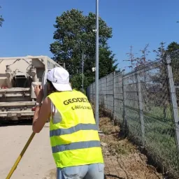 Osoba w kamizelce odblaskowej z napisem 'GEODET' i numerem telefonu, używa niwelatora optycznego na tle ogrodzenia z siatki, zaparkowanego samochodu ciężarowego z zabudową i terenu budowy.