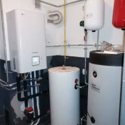 Kotłownia z zamontowaną pompą ciepła LG Therma V, zbiornikami buforowymi i instalacją hydrauliczną w pomieszczeniu z częściowo pomalowanymi ścianami.