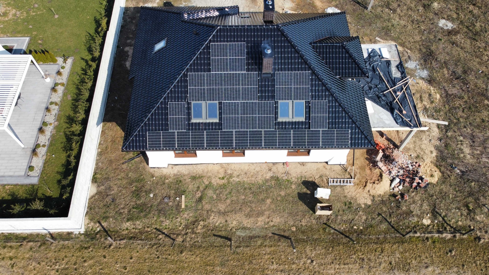Widok z drona na dom z ciemną dachówką, na której zamontowano panele fotowoltaiczne; obok domu widoczne pozostałości po budowie i rozbiórce.