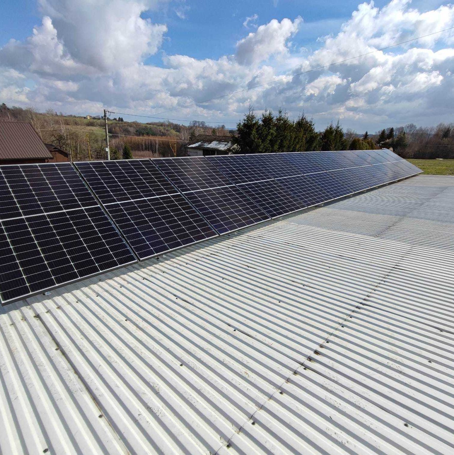Rząd paneli słonecznych na blaszanym dachu z widokiem na pagórkowaty krajobraz i błękitne niebo z chmurami.