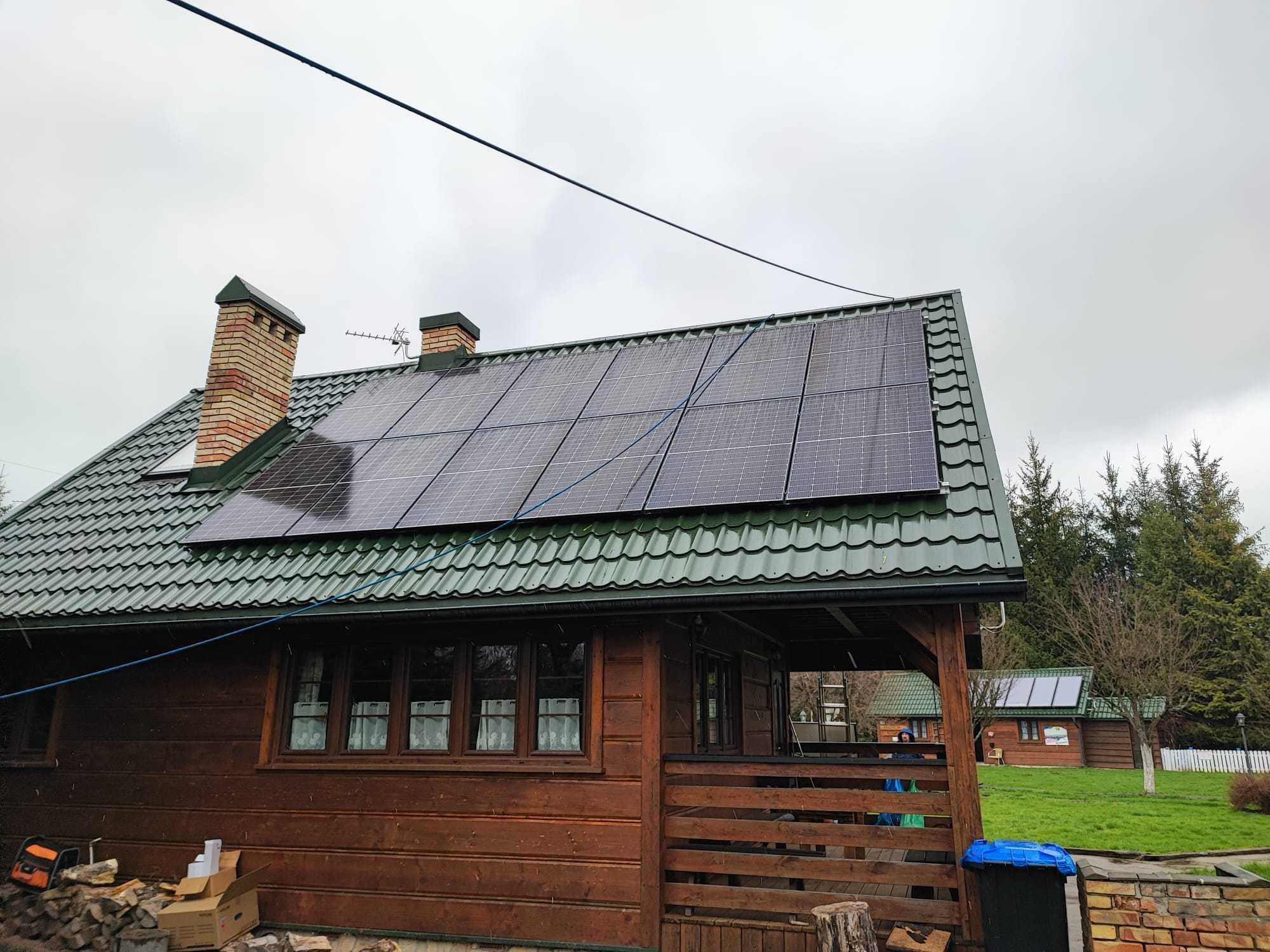Panel słoneczny na dachu drewnianego domu z zieloną dachówką, widoczne kominy z cegły, w tle pochmurne niebo i drugi budynek z panelami.