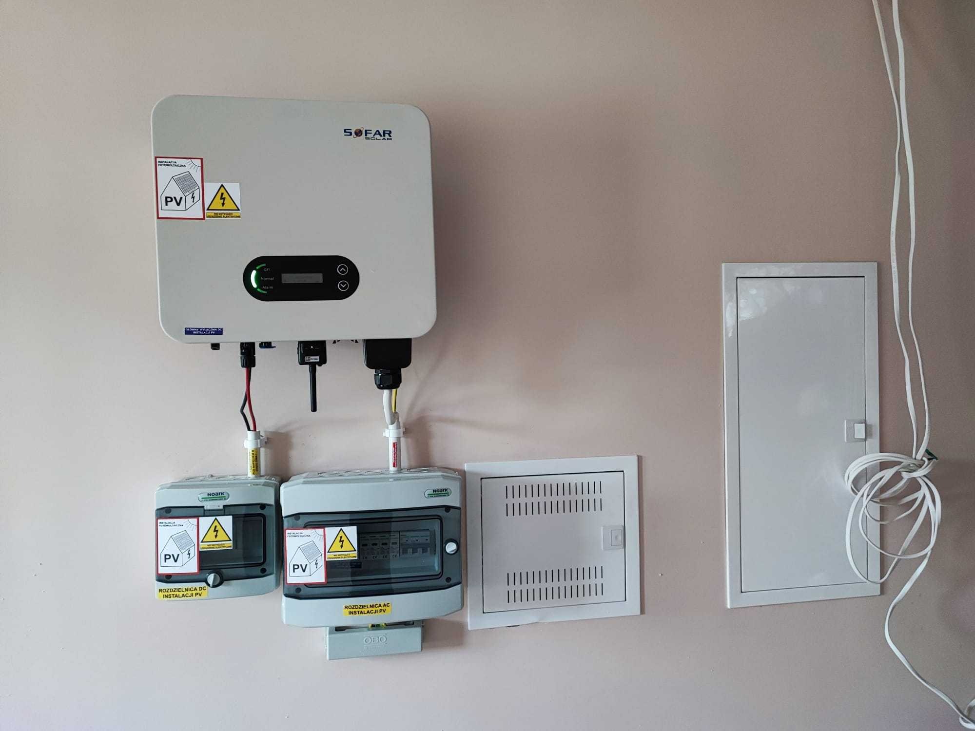 Biały inwerter Sofar Solar z podłączonymi rozdzielnicami DC i AC na ścianie wewnątrz budynku, widoczne naklejki ostrzegawcze i przewody instalacji fotowoltaicznej, obok skrzynki instalacyjne.