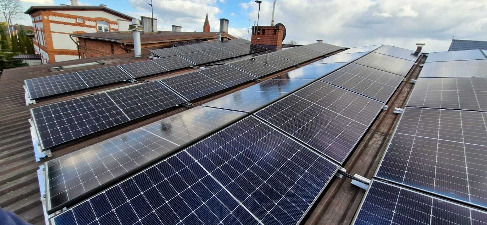 Rzędy paneli słonecznych na falistym dachu budynku mieszkalnego, z odbiciami chmur na powierzchni paneli i widoczną wieżą kościelną w tle.