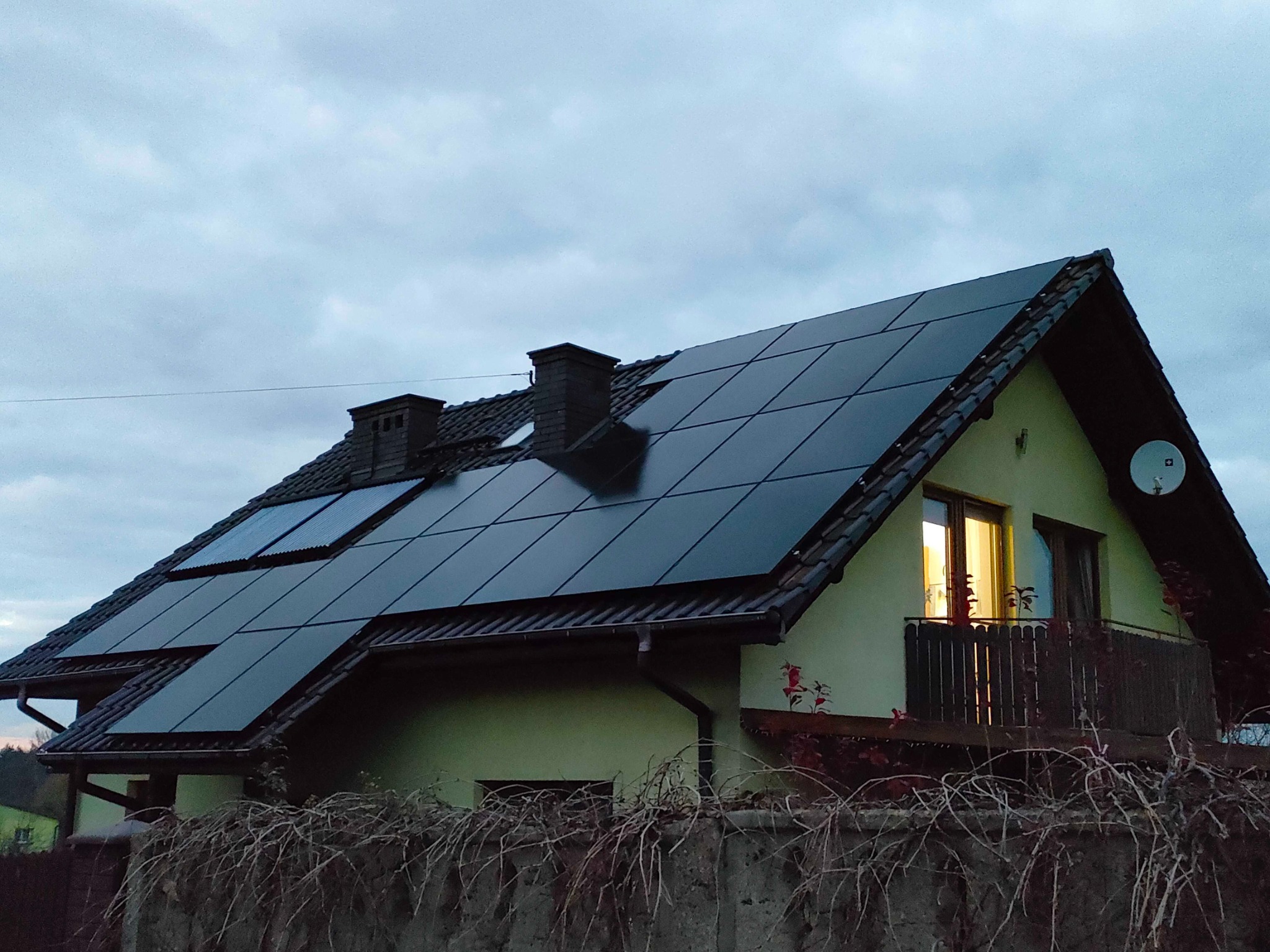 Dom jednorodzinny z panelami fotowoltaicznymi na dachu, widok zza betonowego ogrodzenia z suchymi pnączami, w oknie balkonowym zapalone światło.