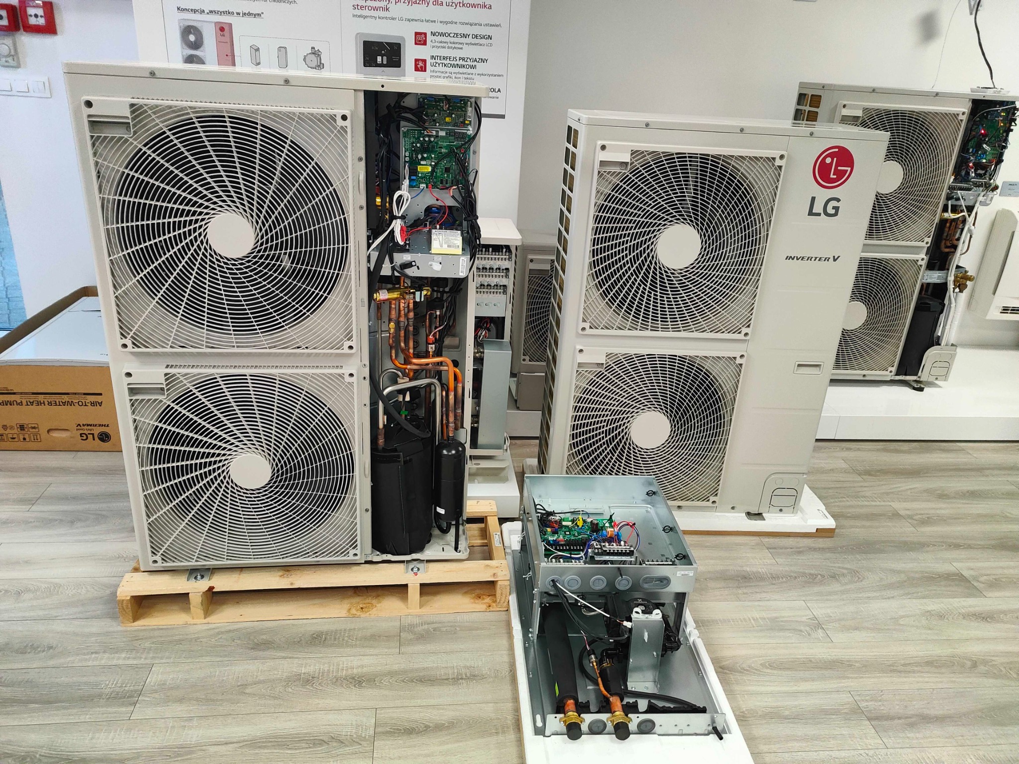 Wnętrze i elementy zewnętrzne klimatyzatorów LG Inverter V w jasnym pomieszczeniu, jeden na palecie, drugi z otwartą obudową ukazującą elektronikę.