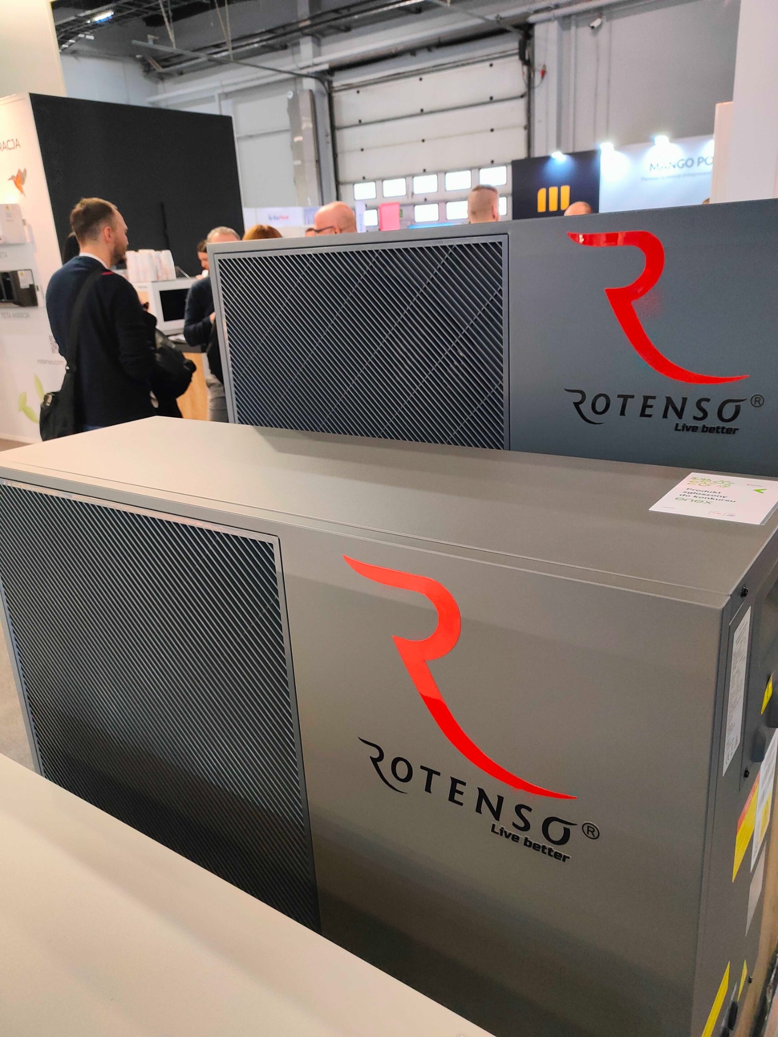Dwa szare urządzenia Rotenso z czerwonym logo i czarnymi kratkami wentylacyjnymi, prezentowane na targach z widocznymi ludźmi w tle.