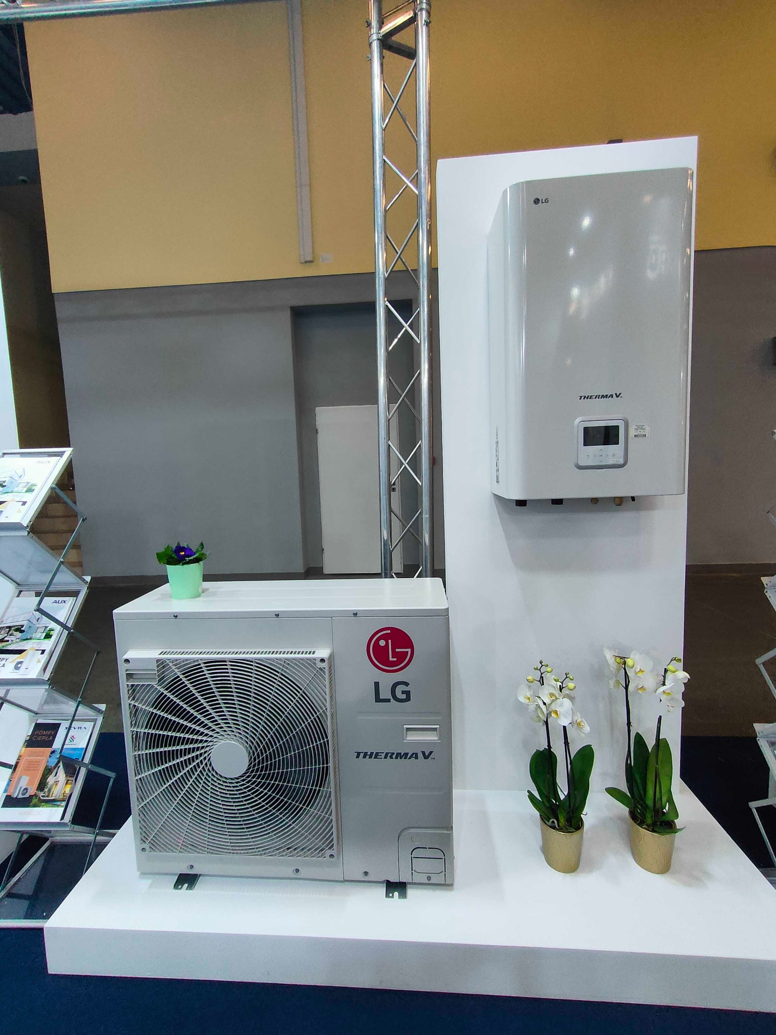 Ekspozycja pompy ciepła LG Therma V na stoisku targowym, z dekoracjami kwiatowymi i fioletowym kwiatkiem doniczkowym na obudowie agregatu zewnętrznego.