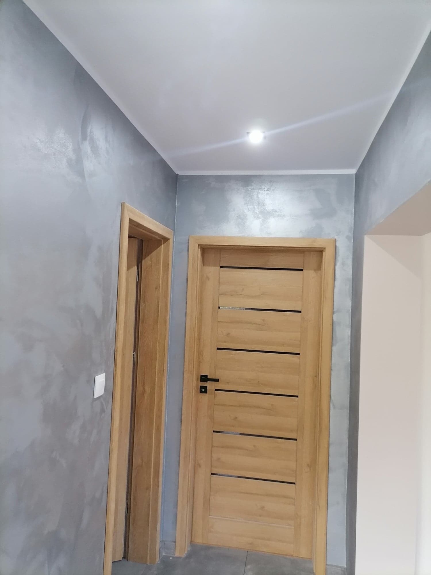 Zamknięte, drewniane drzwi z poziomymi, czarnymi wstawkami w szarej ścianie z efektem dekoracyjnym, widoczny fragment drugich drzwi z ościeżnicą.