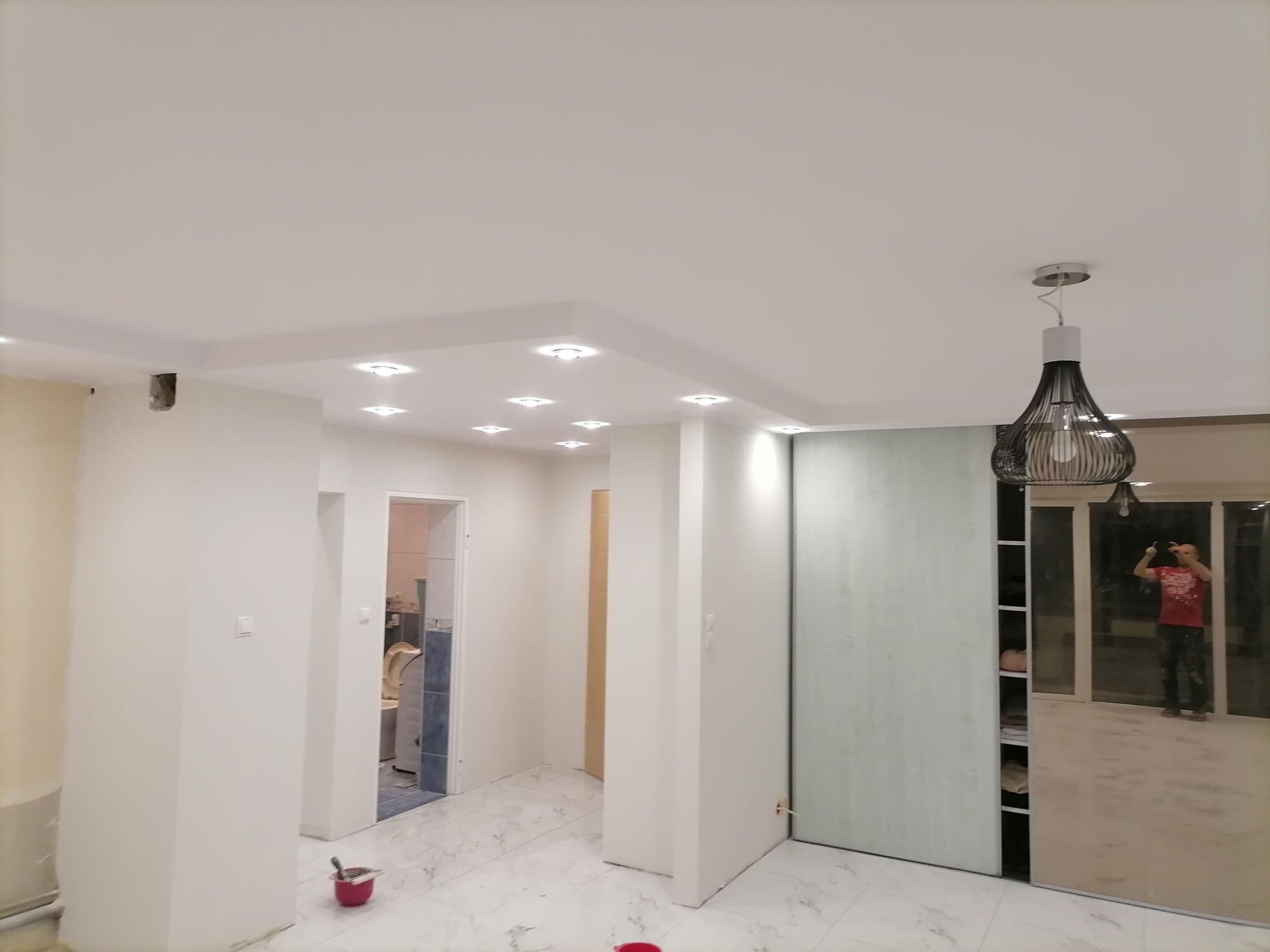 Wnętrze mieszkania w trakcie malowania, widoczne białe ściany, sufit z oświetleniem punktowym, marmurowe płytki podłogowe oraz osoba robiąca zdjęcie w lustrzanej szafie przesuwnej.