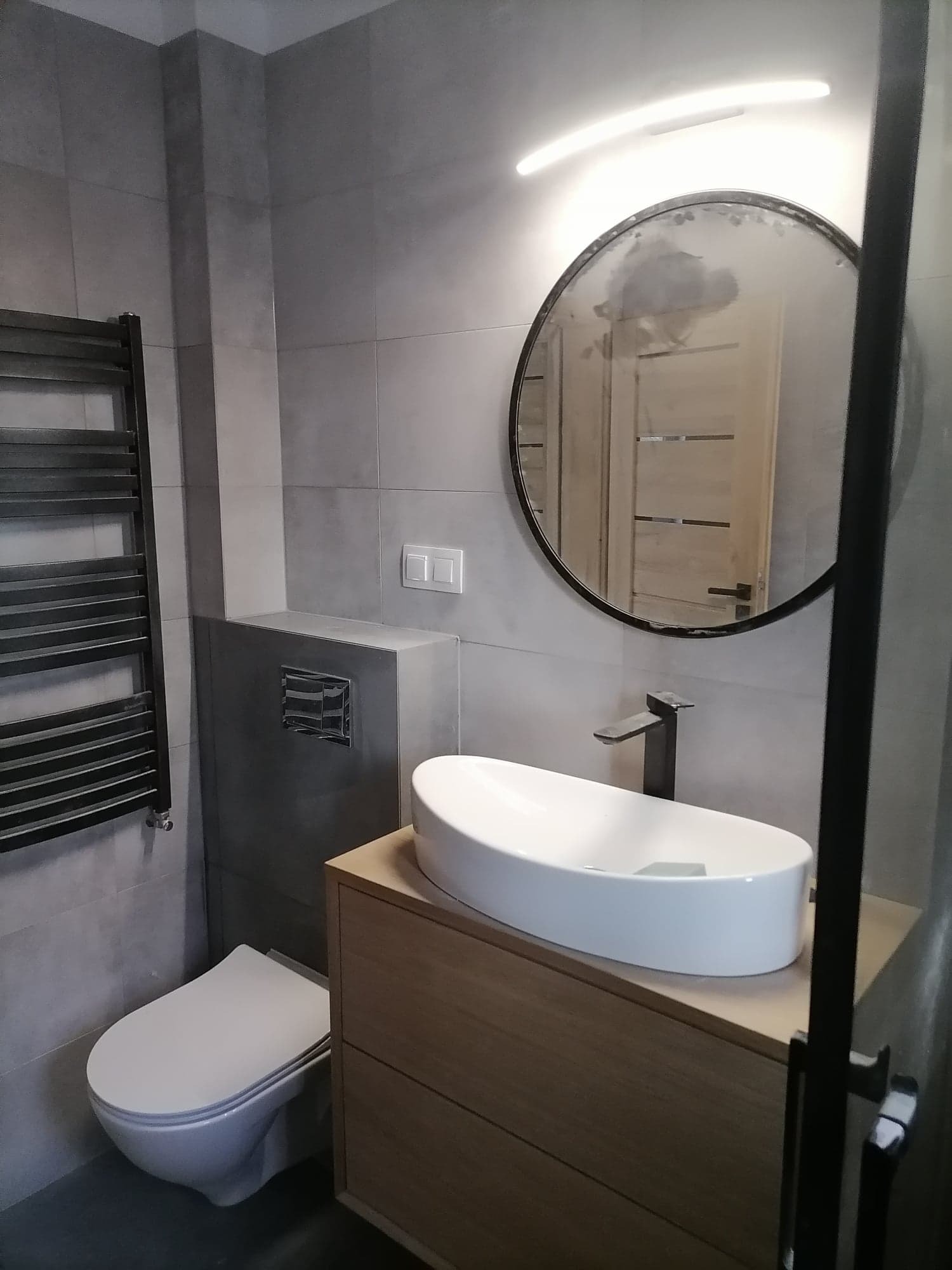 Nowoczesna łazienka w odcieniach szarości z owalną umywalką nablatową, okrągłym lustrem i grzejnikiem drabinkowym.
