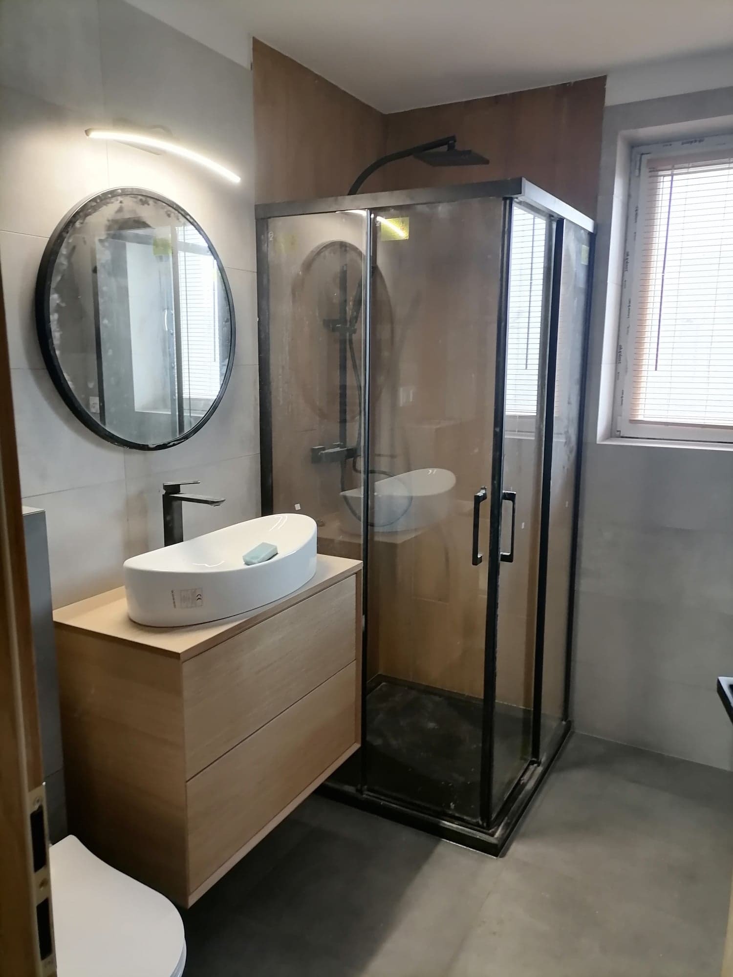 Nowoczesna łazienka z kabiną prysznicową typu walk-in z czarnymi profilami, owalną umywalką nablatową na drewnianej szafce i okrągłym lustrem z podświetleniem LED.