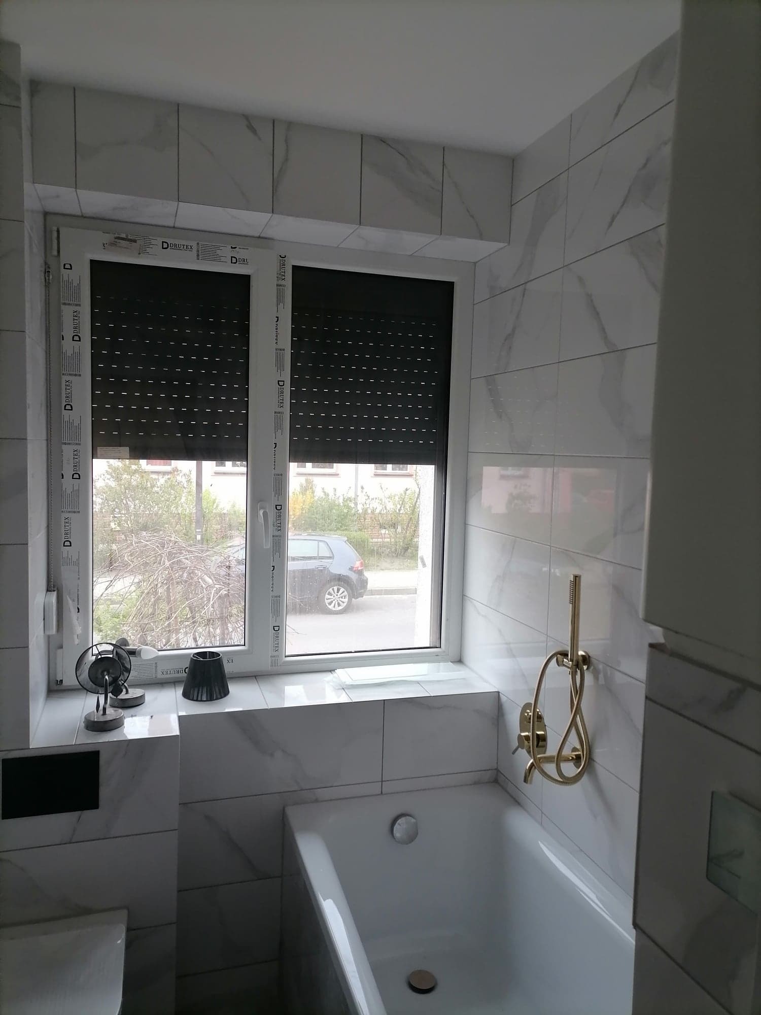 Biała łazienka z wanną, złota bateria prysznicowa, okno z roletami zewnętrznymi i marmurowe płytki na ścianach.