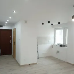 Jasne panele podłogowe w nowym mieszkaniu, białe ściany z punktowym oświetleniem sufitowym i widocznym fragmentem kuchni z oknem.