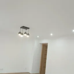 Pomalowane na biało wnętrze z drewnianą podłogą i drzwiami, oświetlone lampą wiszącą z pięcioma żarówkami i punktowymi lampami sufitowymi.