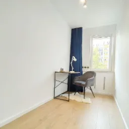 Jasne wnętrze pokoju z panelami podłogowymi, biurkiem z metalową konstrukcją, szarym krzesłem i granatową zasłoną przy oknie z widokiem na budynek.