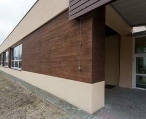 Elewacja budynku z brązowymi panelami imitującymi drewno, widoczne okna i wejście z przeszklonymi drzwiami, ujęcie z perspektywy niskiego kąta.